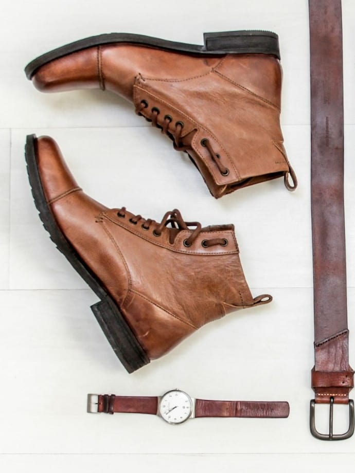 Brown leather boots, a leather belt and a watch laid out on a white floor